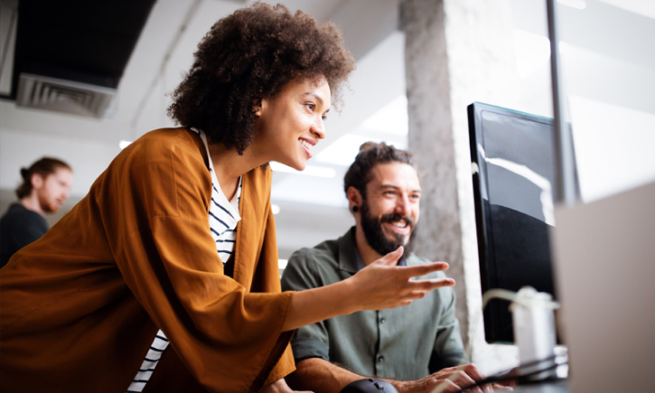 A young woman and a young man look at a computer screen as the woman gestures at it.