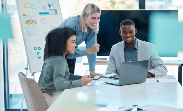 Three young people smile as they gather around a table and look at a laptop together.